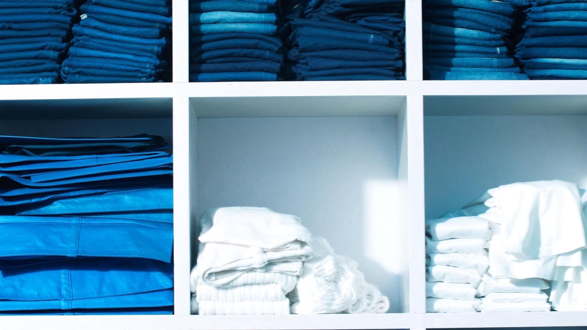 Shelves of folded denim, white knits, and blue-toned fabrics in a bright studio